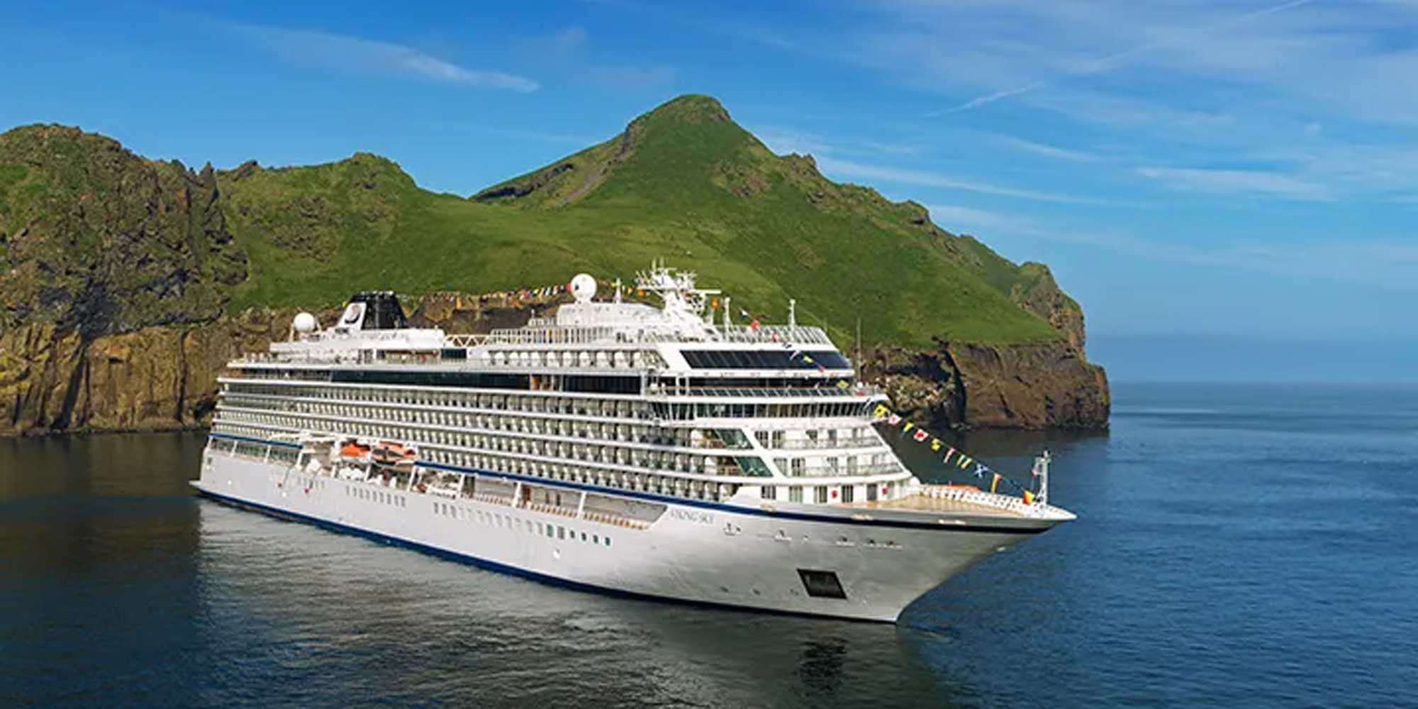 Cruise Ship in Scenic Volcanic Landscape A white luxury cruise ship sailing near a green mountainous island with rocky cliffs and a calm blue ocean. The vessel is adorned with colorful flags and positioned against a picturesque volcanic landscape.