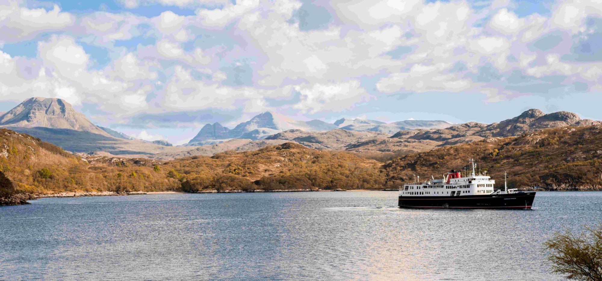 Cruise Ship in Scottish Highlands Waters A classic expedition cruise ship anchors in calm blue waters with dramatic Scottish Highland mountains in the background, including distinctive peaked summits. The rugged landscape features autumn-colored vegetation and dense forests along the shoreline, showcasing the natural beauty of Scotland's remote regions.