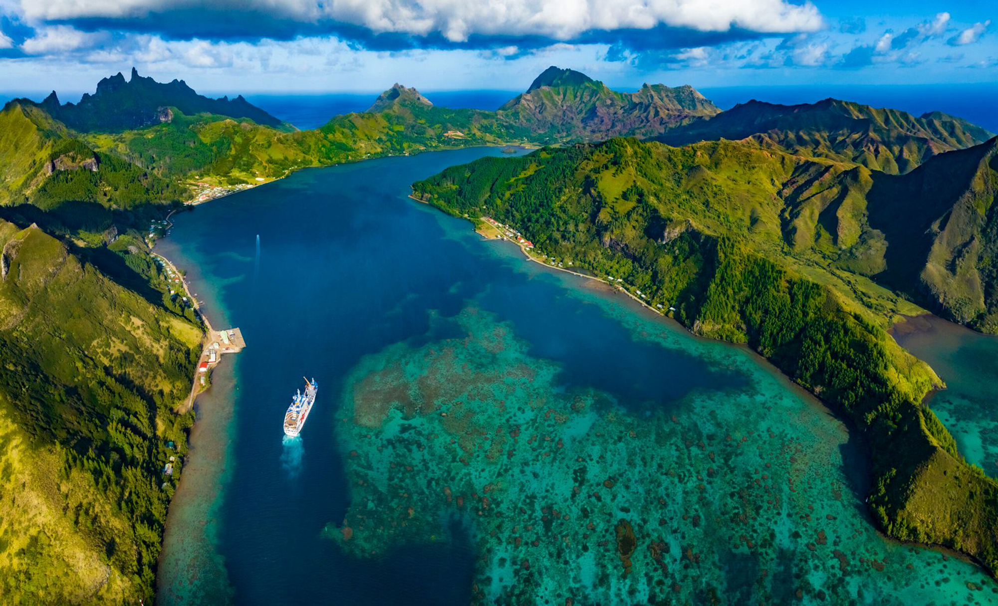 Cruise Ship in Tropical Mountain Lagoon An aerial view of a cruise ship navigating through a stunning turquoise lagoon surrounded by lush green mountainous islands. The landscape features dramatic volcanic peaks and coral reefs visible beneath the crystal-clear waters.
