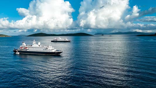 Cruise Ships on Calm Blue Waters Two white cruise ships anchored in a serene blue ocean with dramatic cloudy sky and mountainous islands in the background. The vessels appear to be mid-sized expedition or luxury cruise ships.