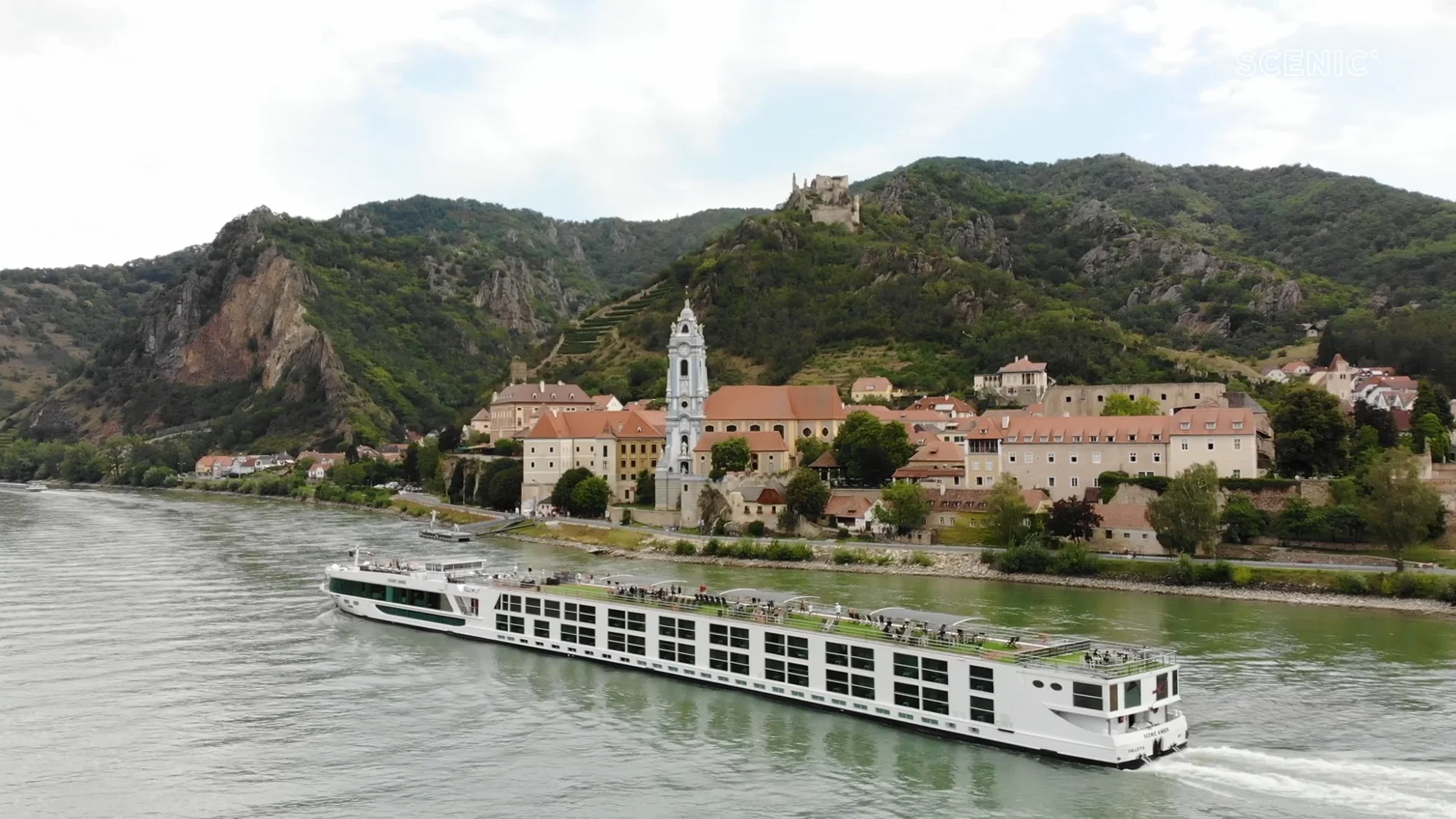 Danube River Cruise in Historic European Landscape A white river cruise ship sailing past a picturesque European town with historic buildings and a church tower, nestled among forested mountains with a castle in the background.