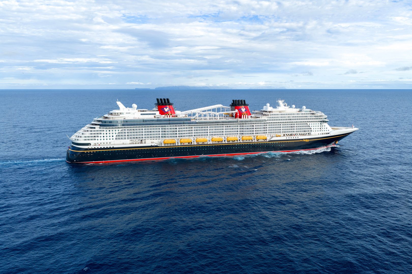 Disney Cruise Line Ship at Sea A massive contemporary cruise ship navigates open ocean waters under a partly cloudy sky. The vessel features distinctive red funnels with black tops, white superstructure, and a dark blue hull with red accent stripe, carrying multiple lifeboats and modern amenities typical of mega-ship cruise lines.