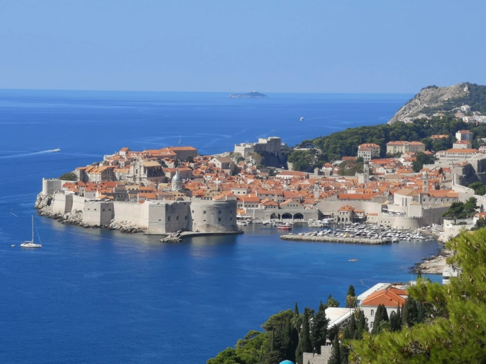 Dubrovnik Old Town Aerial Coastal View A stunning aerial photograph of Dubrovnik's iconic walled Old Town surrounded by brilliant turquoise Adriatic waters, featuring distinctive terracotta-roofed buildings, fortress walls, and the picturesque harbor with moored sailboats. The medieval city is nestled against lush green hillsides with the sparkling blue sea stretching to the horizon.