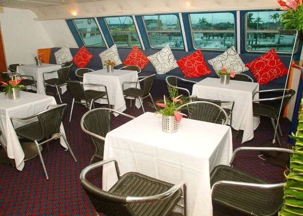 A sophisticated cruise ship dining venue featuring round tables with white linens, woven rattan chairs, and red decorative pillows, overlooking a scenic waterway through expansive windows. The elegant space showcases refined cruise hospitality with tropical floral arrangements and formal table settings.