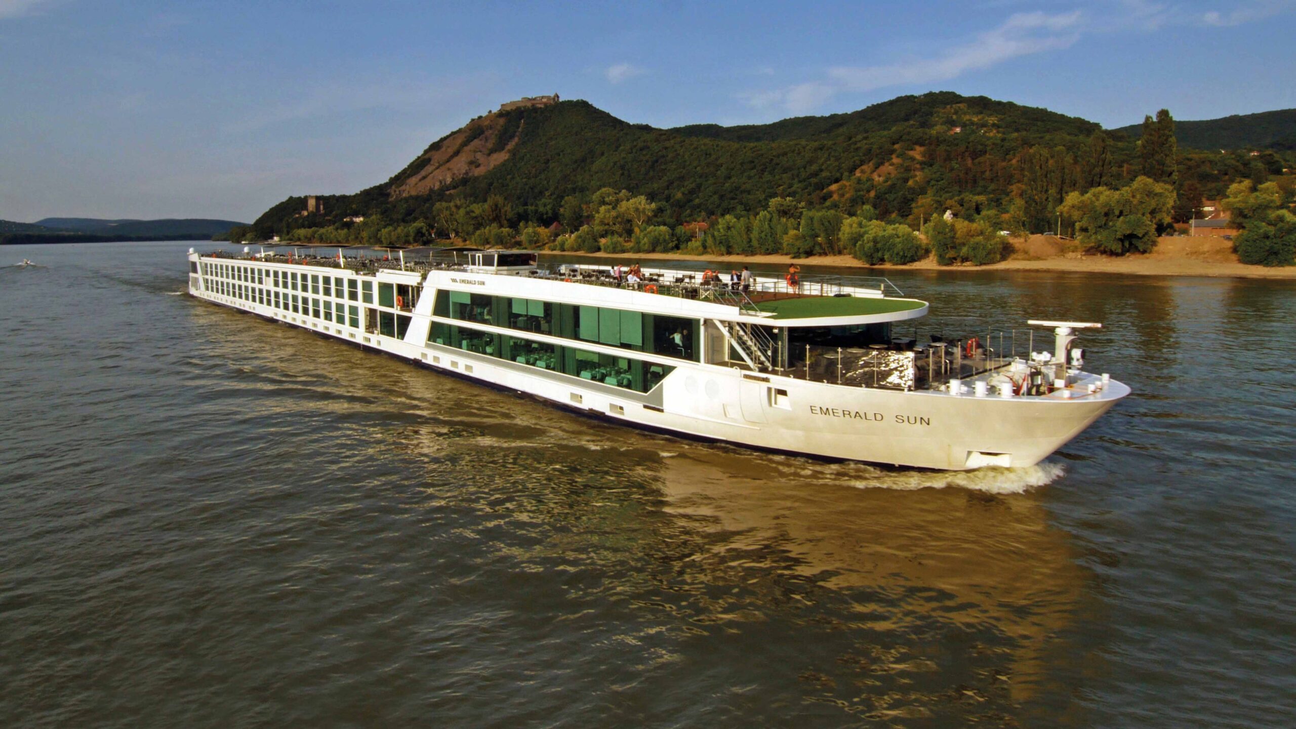 Emerald Sun River Cruise on Scenic Waterway A river cruise ship navigates through a lush, forested river valley with steep hillsides, passing picturesque towns and winding roads along the riverbanks.