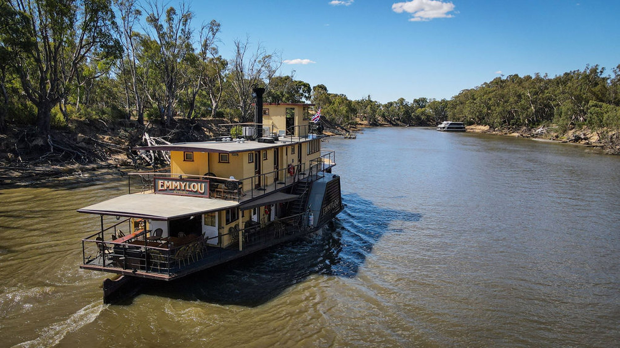 Emmylou Riverboat on Murray River A charming paddle-wheeler riverboat named Emmylou moored along the Murray River in Australia, featuring a distinctive yellow and cream-colored two-story design with open deck areas. The vessel is anchored in shallow water surrounded by native eucalyptus trees and arid landscape under clear blue skies.