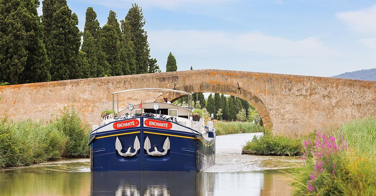 Enchante Barge Passing Under Stone Bridge The MS Enchante, a luxury river cruise barge, navigates through a picturesque European canal passage beneath an ancient arched stone bridge, surrounded by verdant cypress trees and blooming wildflowers along the waterway.