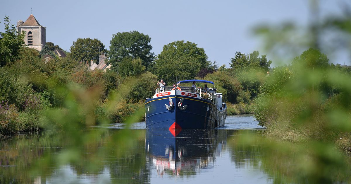 European River Cruise Barge on Waterway A blue and black river cruise barge navigates a peaceful inland waterway lined with lush green vegetation, with a charming church featuring a distinctive bell tower visible in the background among trees. This scene captures the serene experience of river cruising through the European countryside.