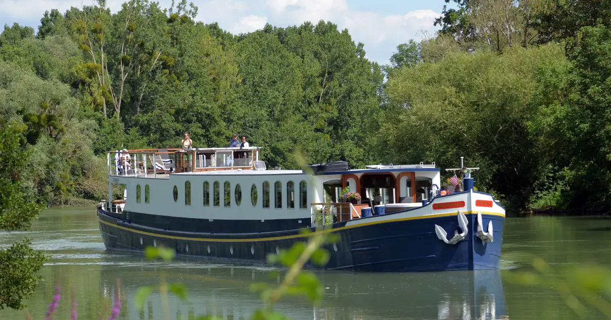 European River Cruise Boat on Waterway A charming European river cruise barge with distinctive blue and white hull navigates a calm waterway surrounded by lush green trees and vegetation. The multi-deck vessel features passenger decks with large windows, an open-air upper deck, and traditional barge styling typical of river cruises along European waterways.