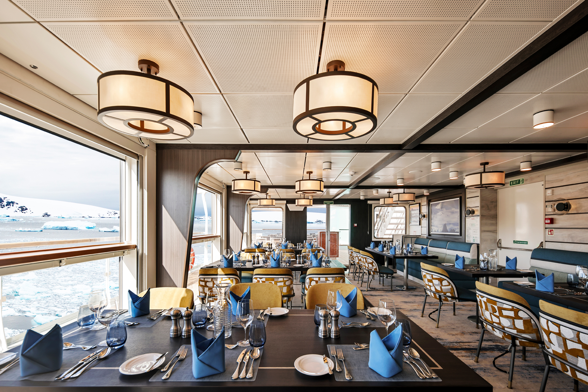 Expedition Cruise Dining Room with Polar Landscape An elegant dining area on an expedition cruise ship featuring large windows overlooking an icy polar landscape with blue-white water and distant snow-covered terrain. The interior has sophisticated lighting, carefully set tables with blue napkins, and yellow accent chairs.