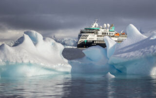 A white and teal expedition cruise ship navigating through blue-white icebergs in a misty polar seascape with dramatic cloudy skies.