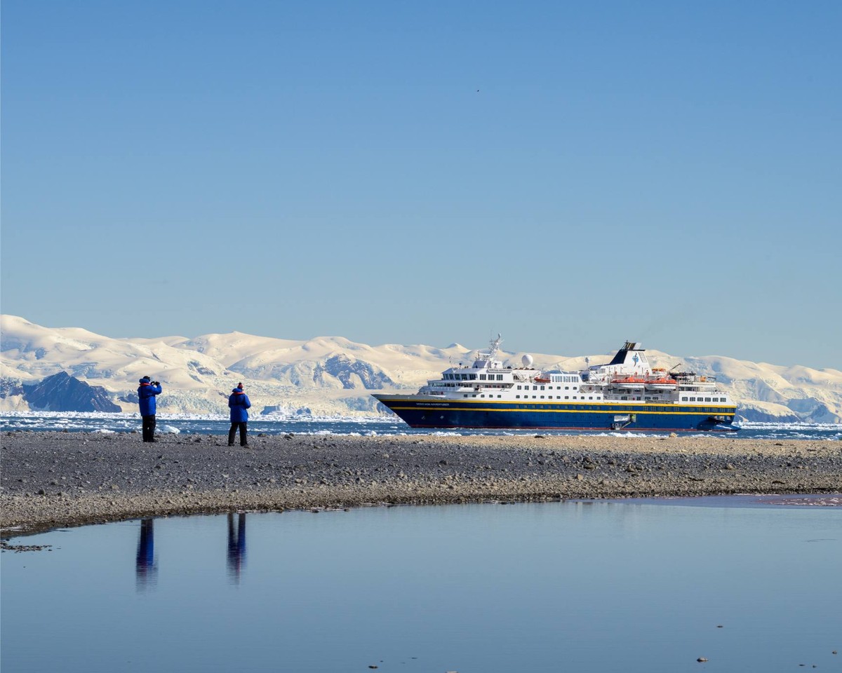 Expedition Cruise Ship in Antarctic Waters A large expedition cruise ship is anchored in pristine Antarctic waters, with passengers on the rocky beach observing the vessel and snow-covered mountains in the background. The clear blue sky and calm waters reflect the ship's distinctive blue and white hull, showcasing the unique polar expedition experience.