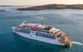 A white expedition-style cruise ship sailing through blue waters near a rocky, forested coastline with small islands in the background. The vessel appears to be designed for adventure and exploration.