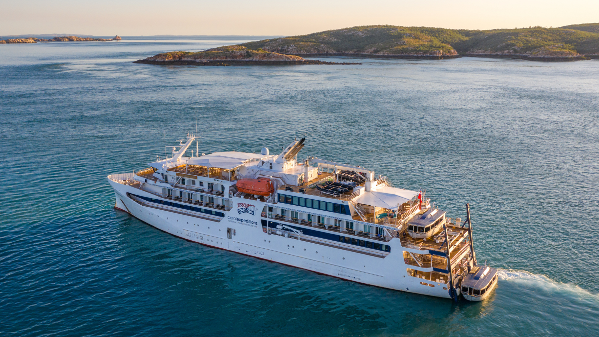 A white expedition-style cruise ship navigating through blue waters near rocky coastal islands during golden hour lighting. The vessel appears to be a smaller, specialized cruise ship designed for adventure travel.