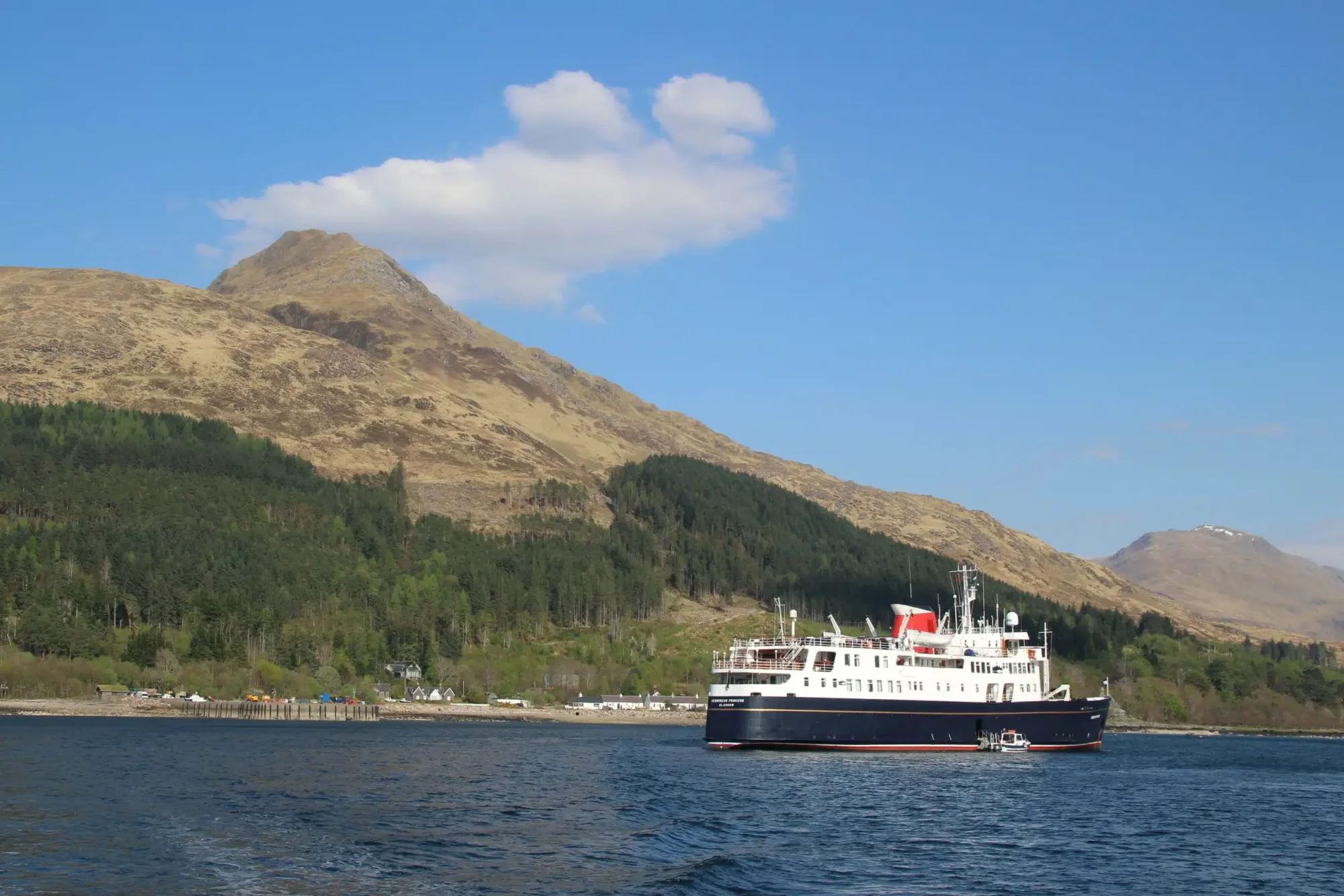 Expedition Cruise Ship in Scottish Highlands A modern expedition cruise ship anchors in a pristine Scottish loch surrounded by dramatic mountainous terrain, dense coniferous forests, and clear blue waters. The vessel, characterized by its white superstructure and red funnel, is positioned against a backdrop of towering peaks and a small shoreside settlement, exemplifying luxury cruise travel in remote wilderness destinations.