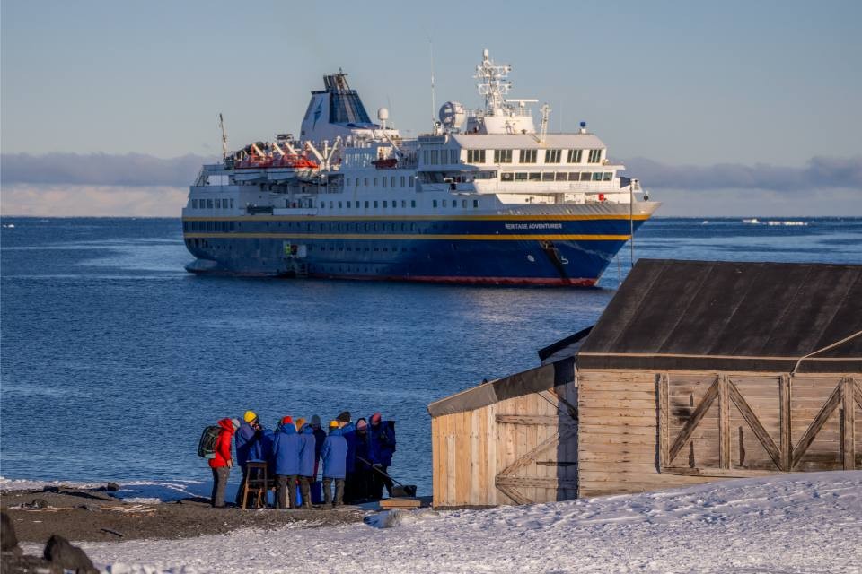 Expedition Ship Arctic Exploration Landing A group of expedition passengers in winter clothing gather on an Arctic beach near a wooden shelter, preparing for or returning from an excursion, with a blue and yellow expedition cruise ship anchored offshore in icy waters. This scene captures authentic polar exploration tourism with the massive vessel serving as a floating base camp for Arctic adventures.