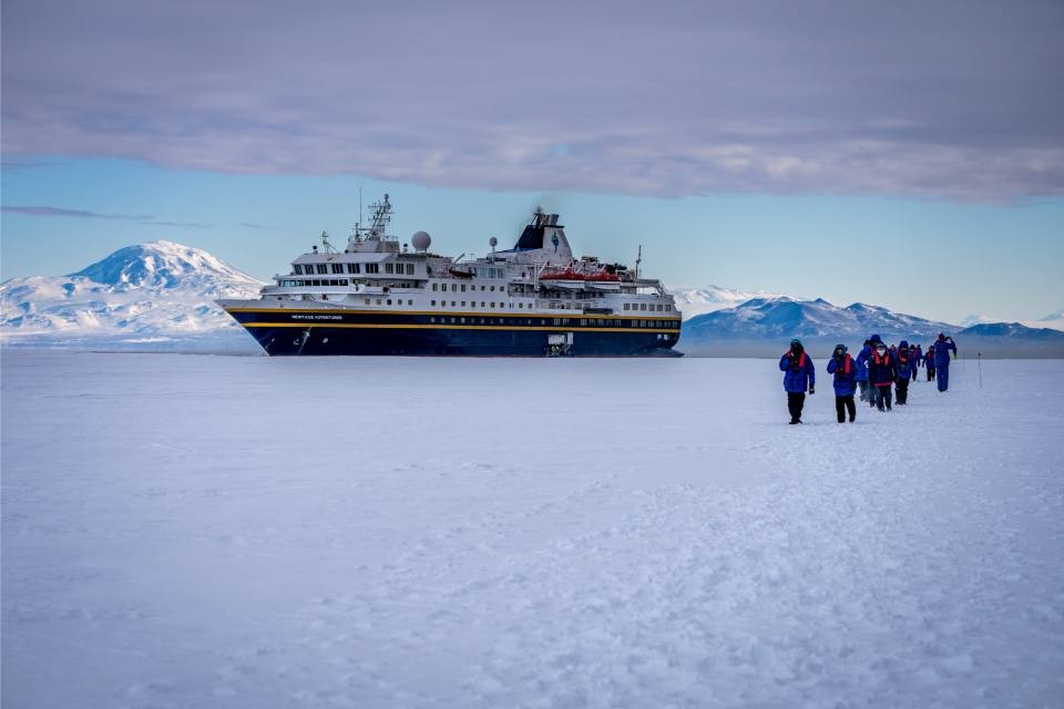 Expedition Ship at Antarctic Ice A large expedition cruise ship anchored in Antarctic waters with passengers walking across the frozen sea ice toward the vessel. Snow-capped mountains are visible in the background under a partly cloudy sky, showcasing polar exploration and adventure tourism in one of Earth's most remote destinations.