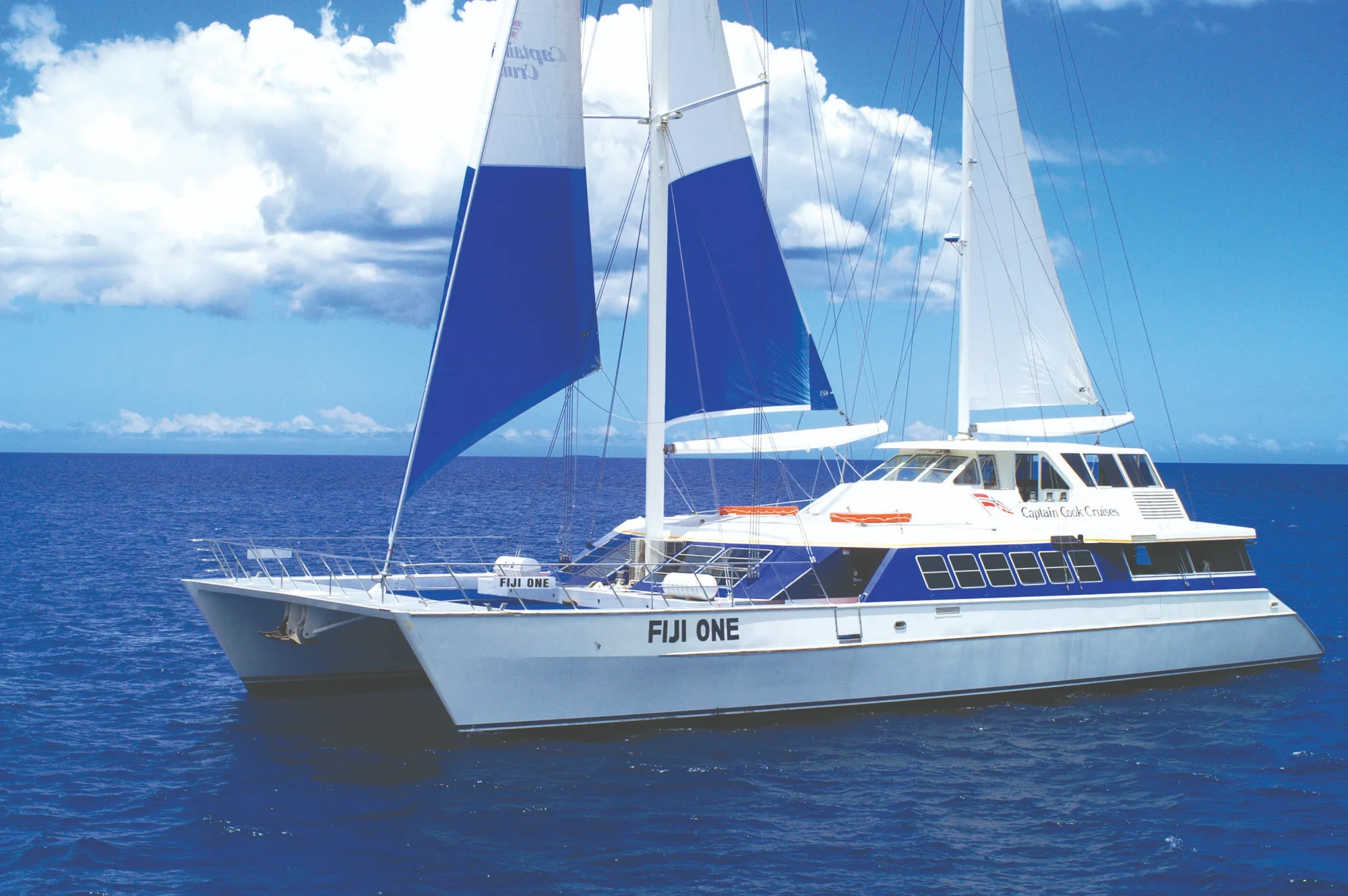 The Fiji One, a modern sailing catamaran operated by Captain Cook Cruises, glides across crystalline blue waters with billowing white and navy sails deployed. The twin-hulled vessel features multiple decks and comfortable outdoor spaces, exemplifying luxury small-ship cruising in tropical destinations.
