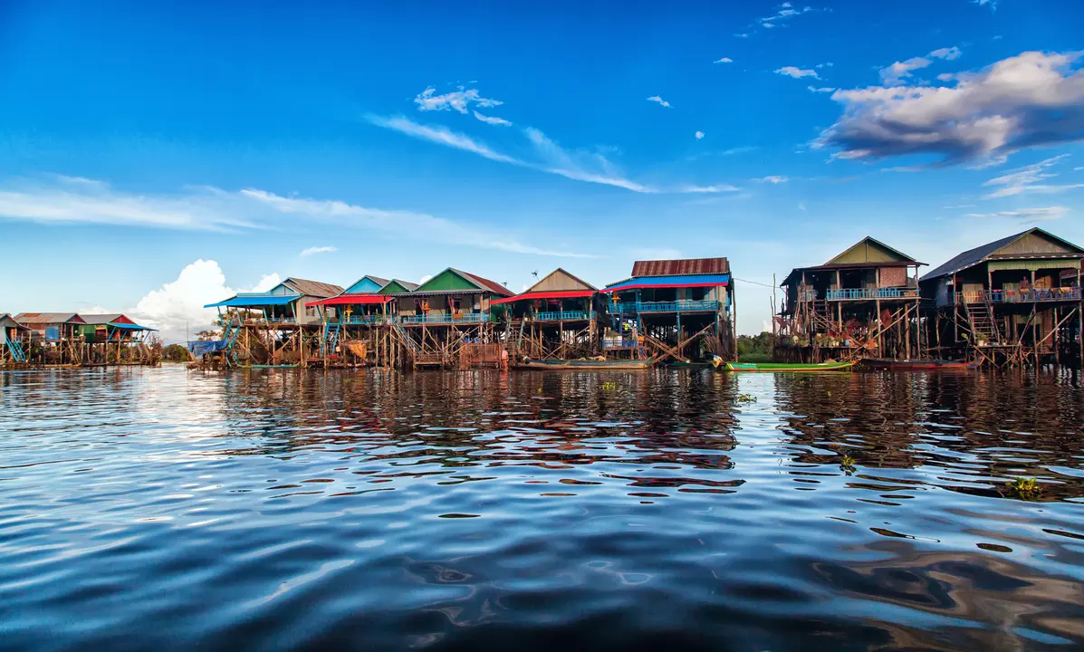 Floating Village on Tonlé Sap Lake A vibrant floating village featuring traditional wooden stilt houses with colorful roofs reflected in calm water, showcasing the unique lifestyle and architecture of communities living on Southeast Asian waterways. The scene captures the authentic cultural experience of Cambodian waterside settlements with clear blue skies and pristine natural surroundings.