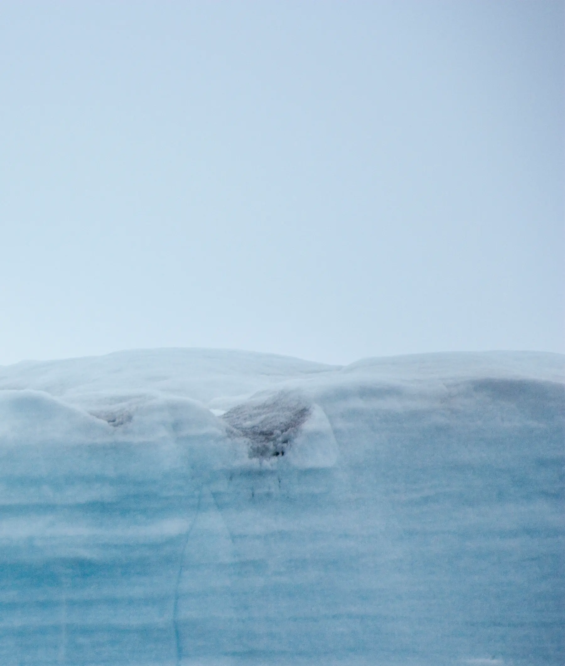 Glacial Ice Formation in Arctic Waters A striking photograph of a massive blue glacier or iceberg with layered ice formations rising above calm Arctic waters, captured under a clear, pale sky that emphasizes the stark beauty and scale of the polar landscape.