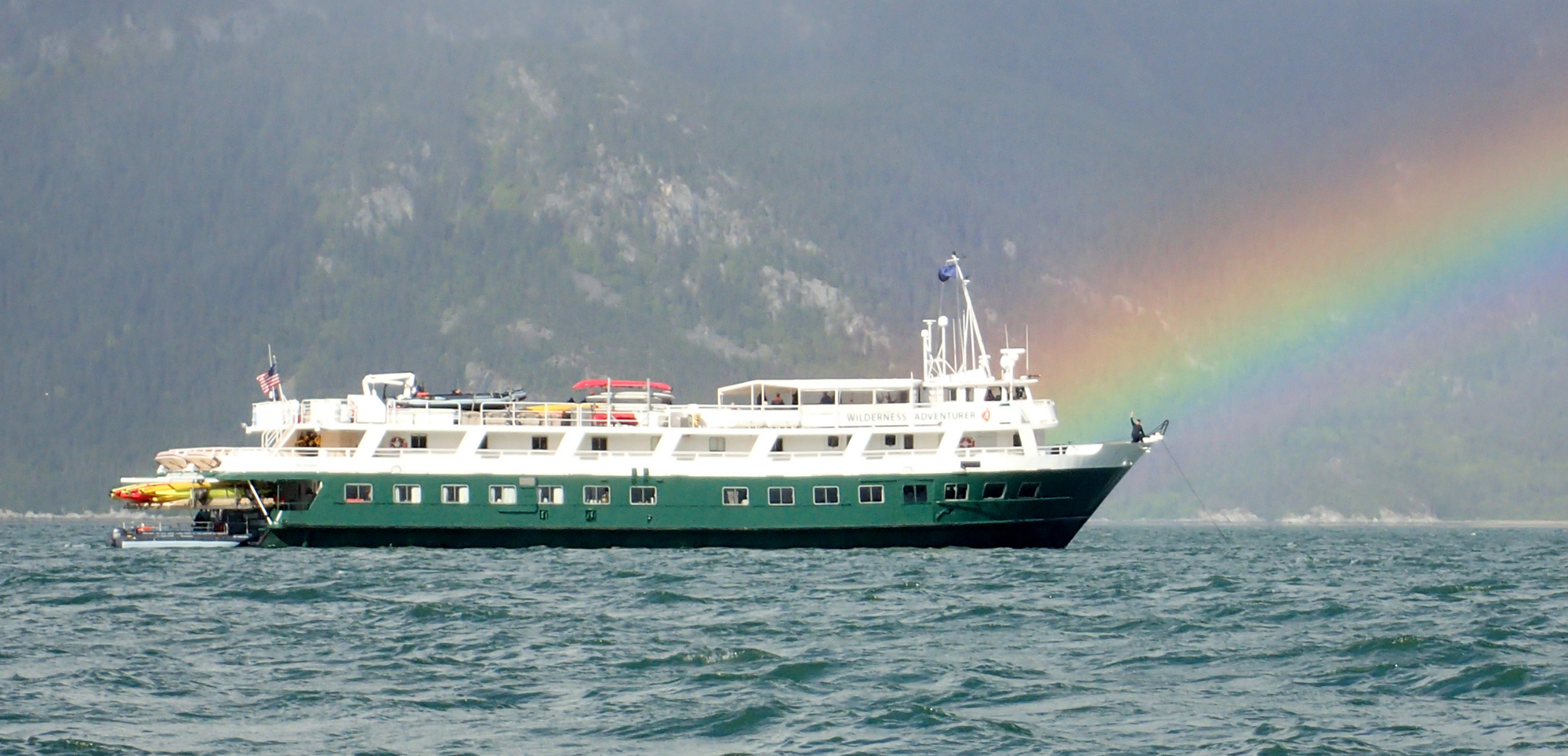 Green Cruise Ship with Rainbow Backdrop A green and white expedition or cruise ship sailing on choppy waters with a vivid rainbow arching in the background and misty mountainous terrain visible.