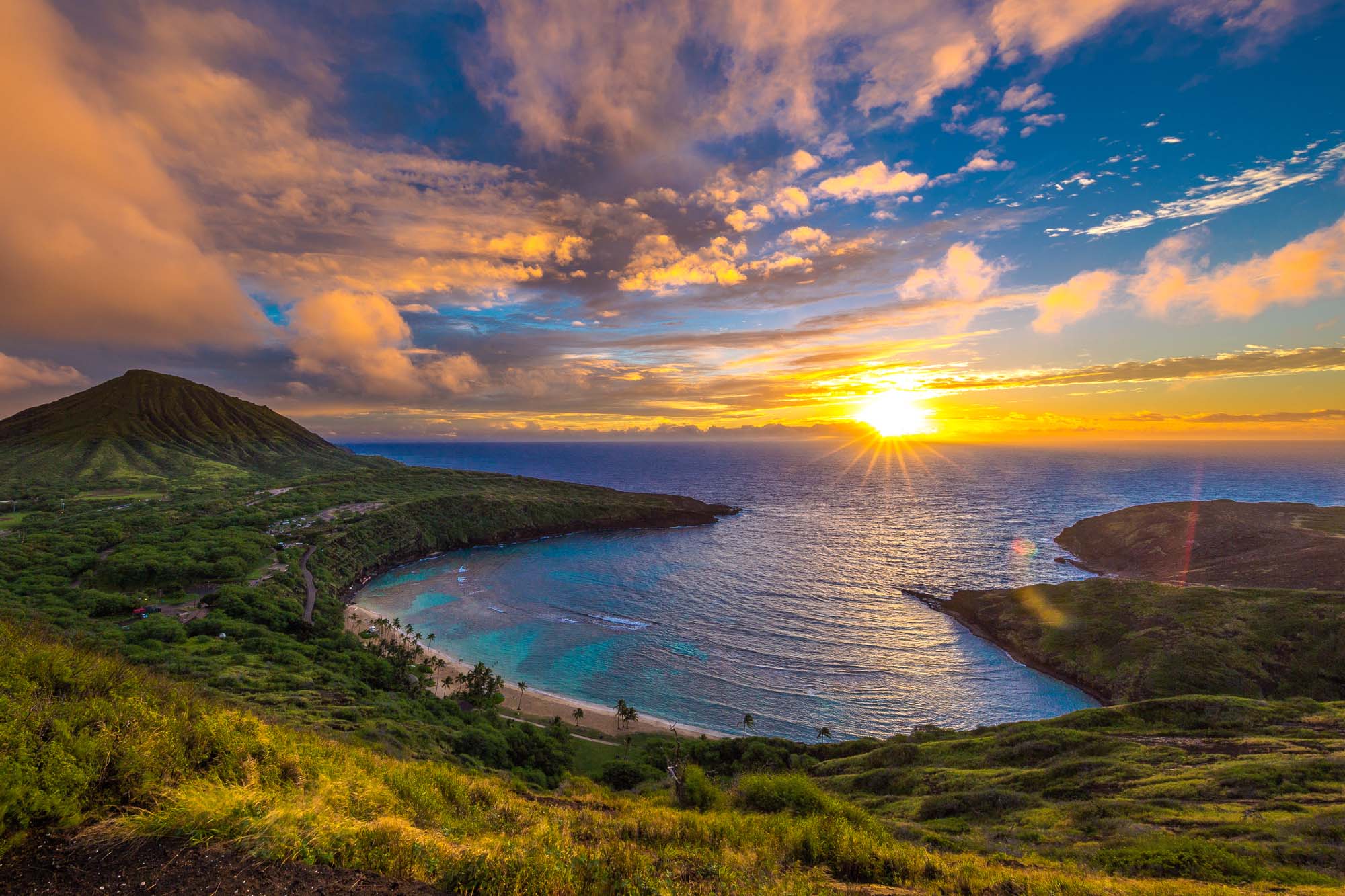 A breathtaking sunrise view over Hanauma Bay on Oahu, Hawaii, featuring a crescent-shaped volcanic cove with pristine turquoise waters, golden sandy beach, lush green hillsides, and dramatic coastal cliffs bathed in warm golden light as the sun rises over the Pacific Ocean.