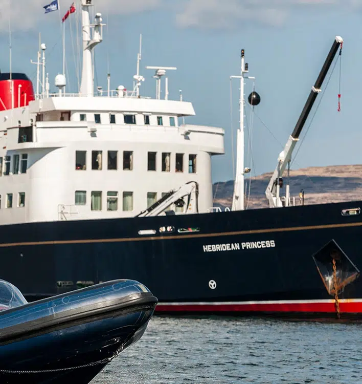 Hebridean Princess Cruise Ship at Port The Hebridean Princess, a small luxury cruise ship, is moored at a Scottish port with its distinctive white superstructure and black hull clearly visible. A small tender boat is secured alongside the vessel, with rolling hills visible in the background under a partly cloudy sky.