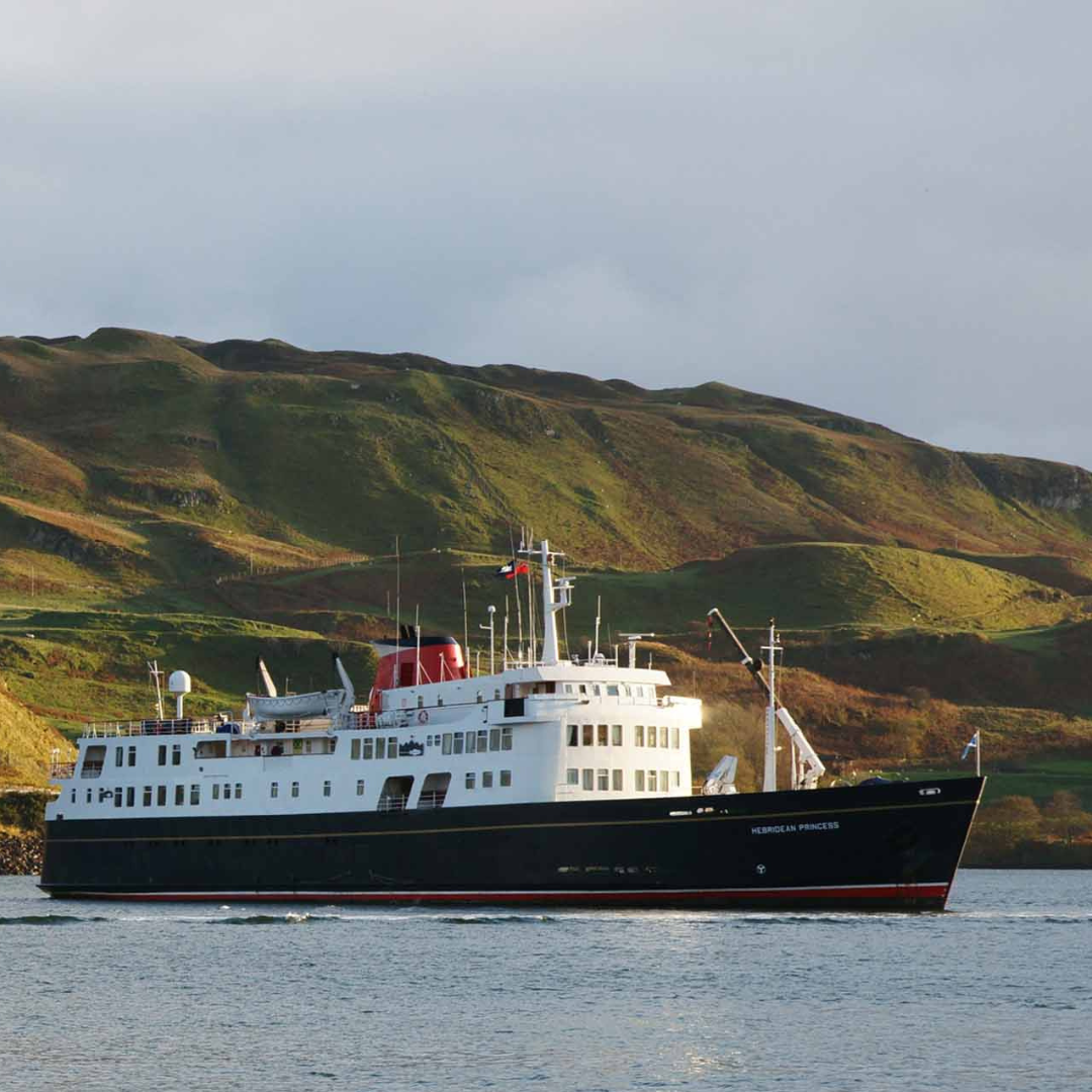 Hebridean Princess in Scottish Waters The Hebridean Princess, a small luxury expedition cruise ship, anchors in calm waters with dramatic moss-covered hills and mountains rising behind it. The vessel features classic white superstructure with a black hull and red accent stripe, displaying flags characteristic of Scottish waters and remote island destinations.