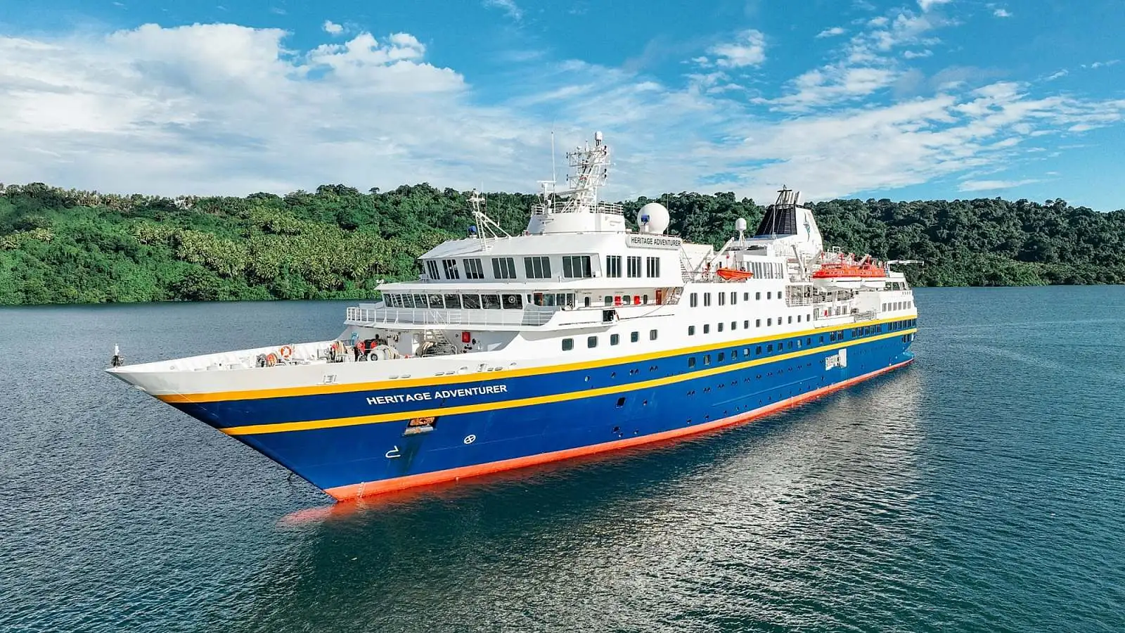 The Heritage Adventurer, a modern expedition cruise ship with distinctive blue and orange hull livery, anchors in turquoise tropical waters surrounded by lush green mangrove forests. The vessel features multiple decks, advanced navigation equipment, and is designed for adventure cruising to remote destinations.