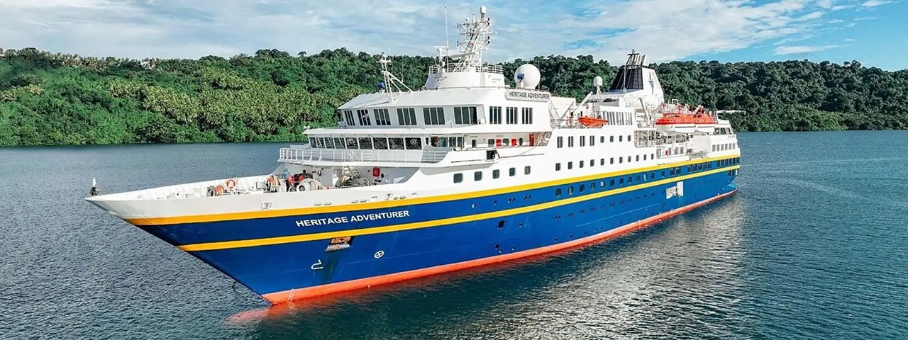 Heritage Adventurer Expedition Cruise Ship The Heritage Adventurer, a modern expedition cruise vessel, anchors in turquoise tropical waters with lush green mangrove forests lining the shoreline. The ship features a distinctive blue hull with yellow stripe, white superstructure, and is equipped with expedition amenities visible on deck.
