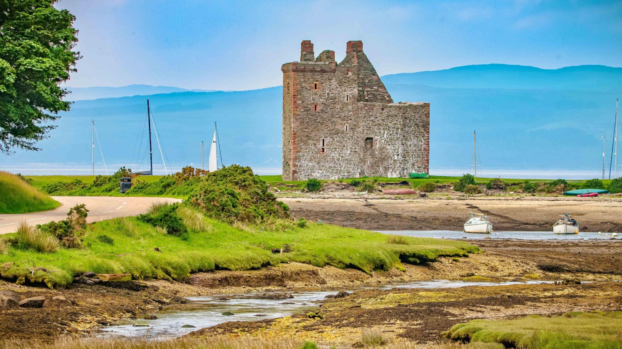 Historic Castle Ruins by Scottish Coastal Waters A picturesque medieval stone castle tower stands on the banks of a Scottish estuary, surrounded by lush green marshland and moored sailboats. The ruins showcase traditional Scottish architecture with mountains visible across the water under a clear blue sky.
