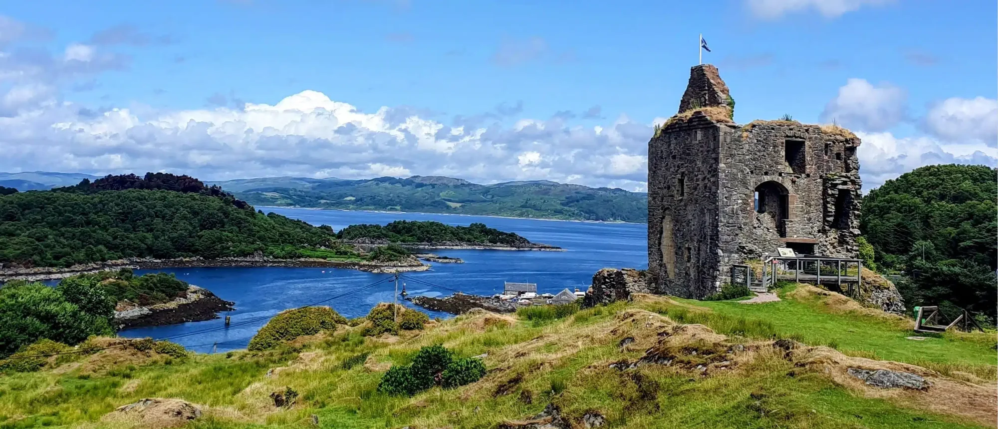 Historic Castle Ruins on Scottish Coastline A dramatic stone castle ruin with a flagpole stands on a green clifftop overlooking a picturesque Scottish coastal landscape, with deep blue waters, forested headlands, and distant mountains beneath a partly cloudy sky.