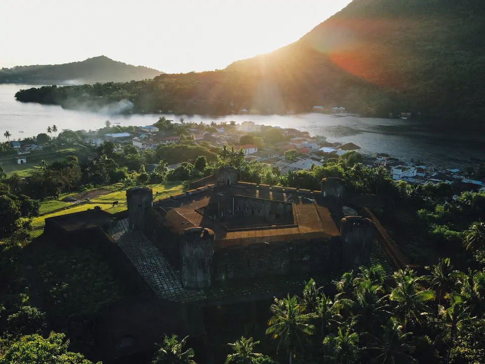 Historic Fort Overlooking Tropical Island Village An aerial view of a weathered colonial-era fortification overlooking a lush tropical island settlement nestled along a misty waterfront at sunrise or sunset. The golden light illuminates the surrounding landscape, revealing verdant vegetation, traditional buildings, and the serene waters beyond, creating a picturesque destination for maritime exploration.