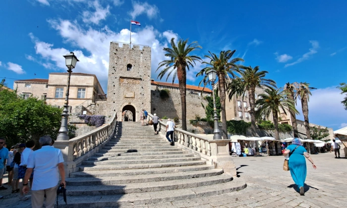 Historic Stone Tower in Mediterranean Coastal Town A scenic plaza featuring a medieval stone tower with a flag at its peak, connected by a grand staircase with ornate white balustrades. Palm trees and historic architecture frame the picturesque European destination under a bright blue sky.