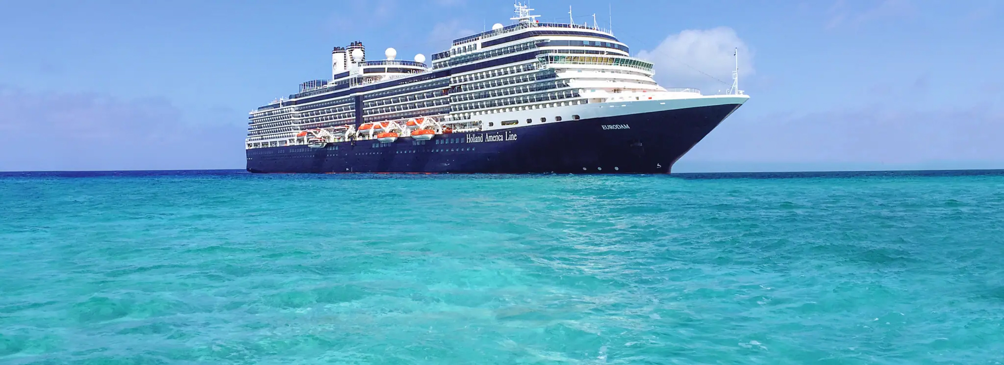 A large blue and white Holland America cruise ship sailing on turquoise Caribbean waters under a clear blue sky. The vessel appears to be a modern, mid-sized cruise liner anchored in pristine tropical waters.