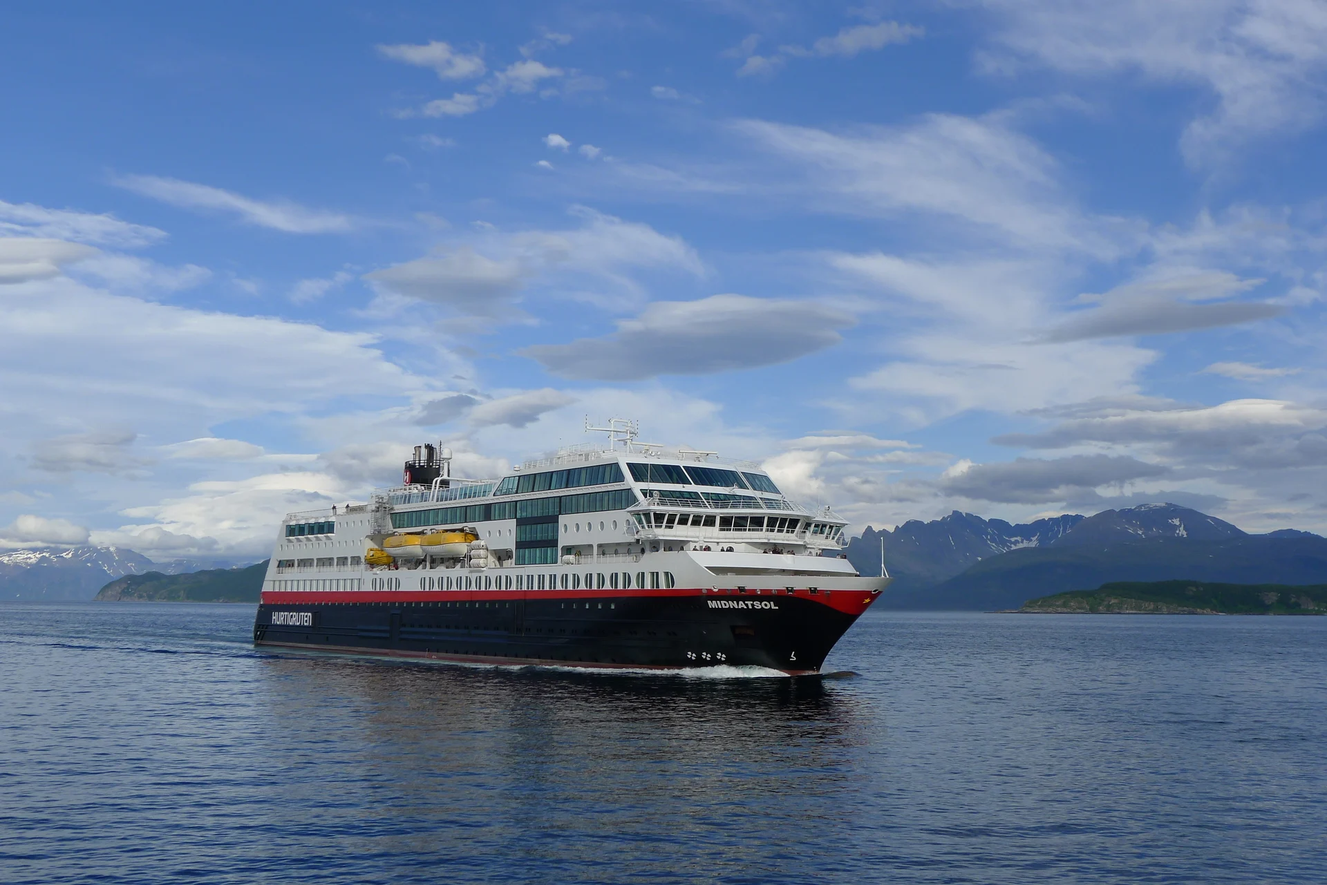 A Hurtigruten cruise ship navigating through a scenic Norwegian fjord landscape with snow-capped mountains in the background under a dramatic cloudy sky.