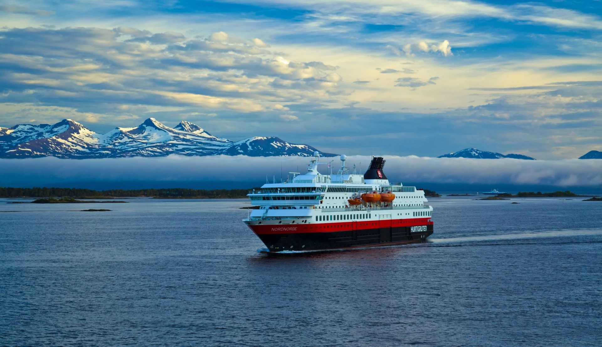 A Hurtigruten cruise ship sailing through a scenic Norwegian fjord landscape, with snow-capped mountains in the background and misty waters surrounding the vessel.