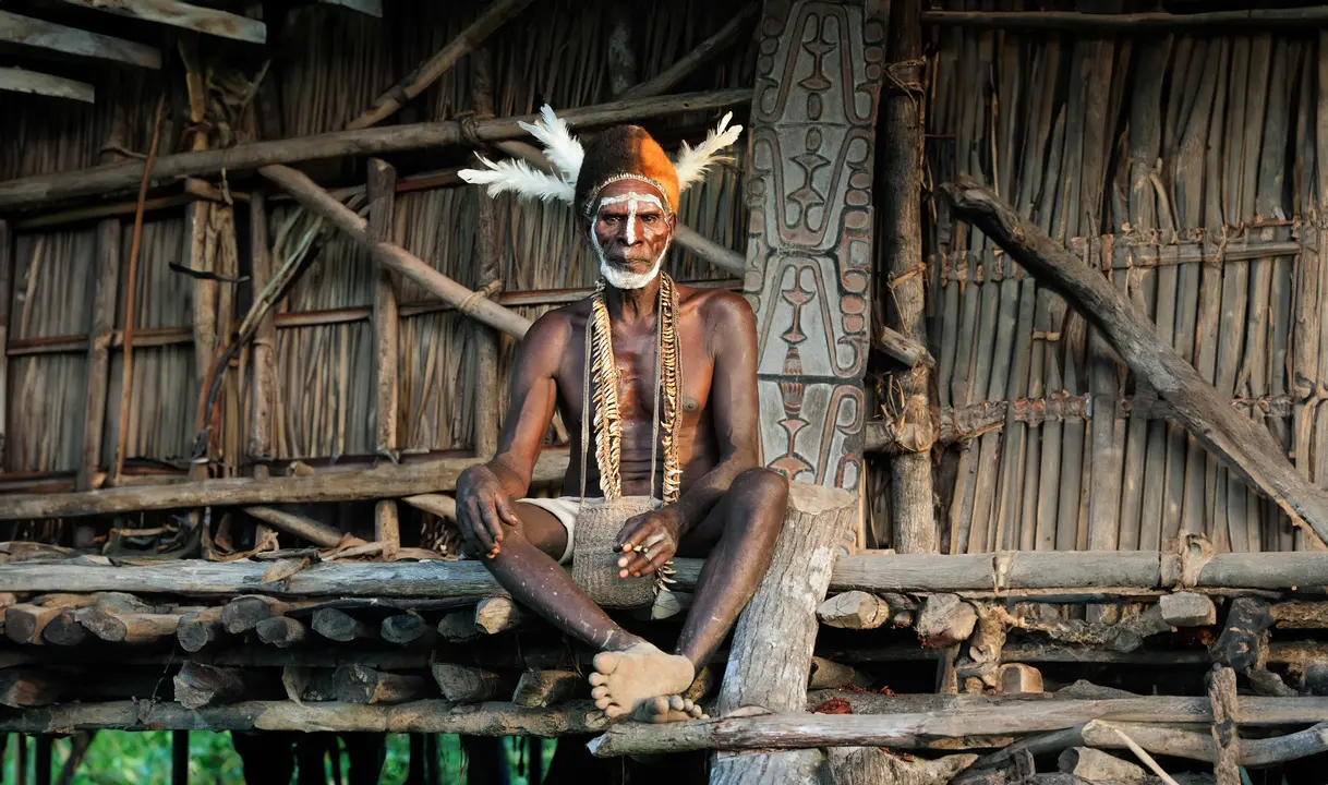 Indigenous Papua New Guinea Tribal Elder A dignified tribal elder from Papua New Guinea sits on a traditional wooden structure, displaying authentic cultural attire including face paint, feathered headdress, beaded necklaces, and woven garments. The background showcases traditional architecture with intricate carved wooden panels characteristic of Papuan indigenous communities.
