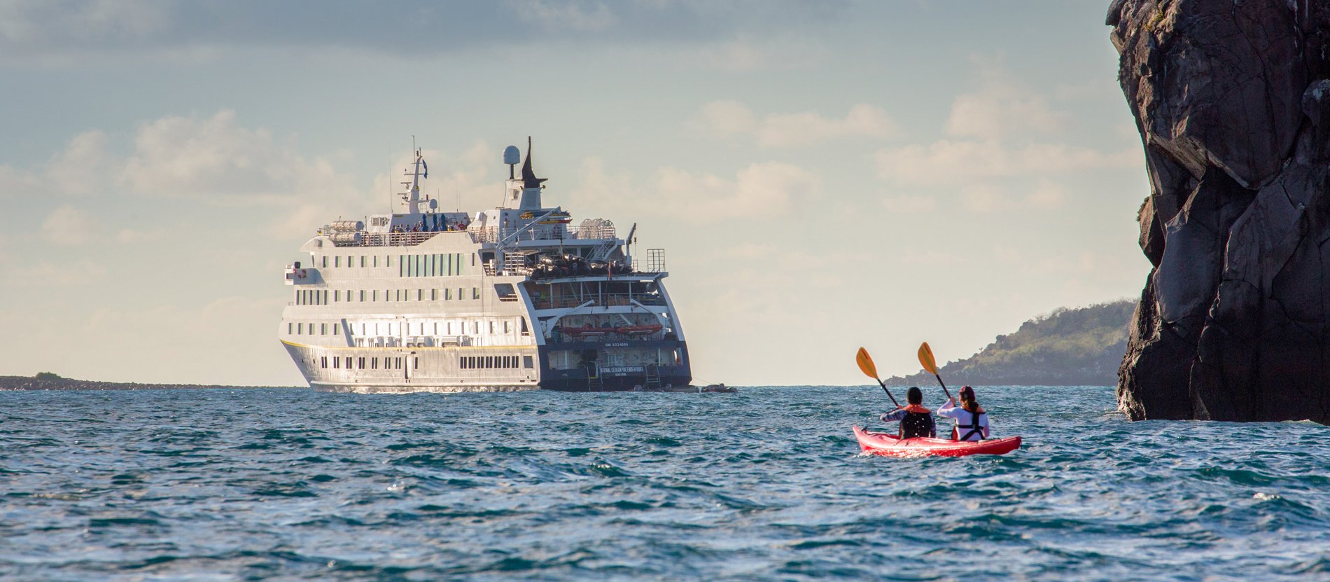 Kayaking Adventure Near Cruise Ship Two kayakers in a red kayak paddle near dramatic limestone cliffs while a large white cruise ship anchors in the turquoise waters beyond. This scene captures the adventure and exploration opportunities available during cruise port stops in tropical destinations.