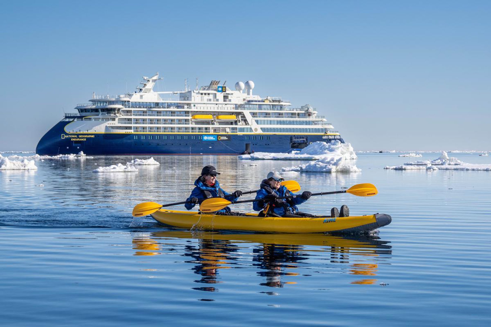 Two kayakers paddle in a bright yellow kayak in pristine Arctic waters surrounded by icebergs, with the National Geographic Endurance cruise ship visible in the background. This scene captures an expedition cruise experience combining adventure activities with luxury polar exploration.