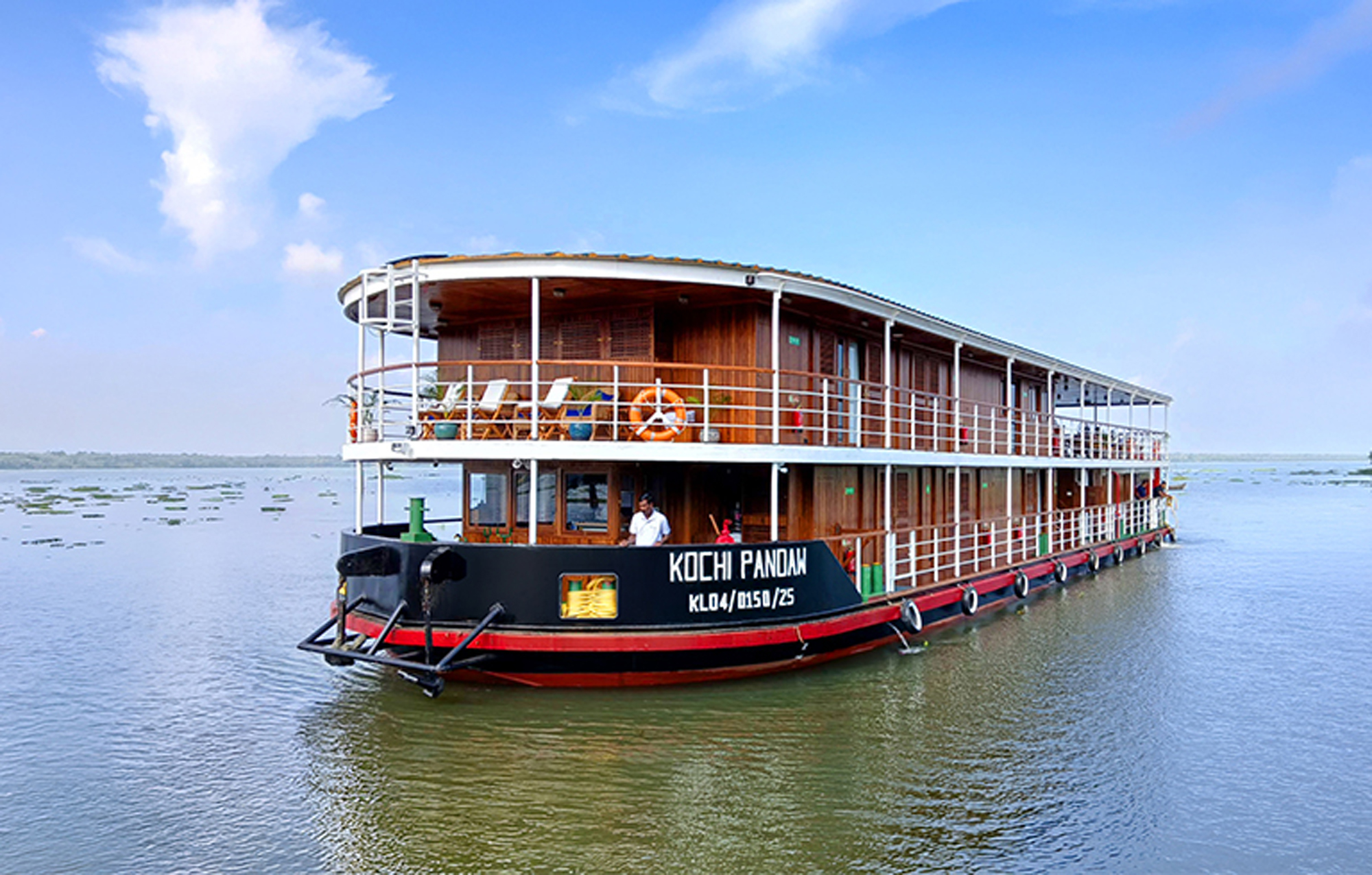 Kochi Pandam Riverboat in Kerala Backwaters A two-story wooden riverboat named Kochi Pandam floating on calm green waters with a blue sky and white clouds in the background. The boat features multiple decks with wooden paneling and white railings.