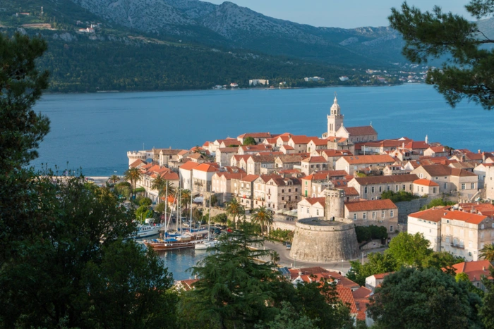 Korcula Old Town Dalmatian Coast Croatia A picturesque medieval walled town on the Dalmatian coast of Croatia, featuring distinctive red-tiled roofs, a prominent church bell tower, and traditional architecture nestled on a peninsula surrounded by azure waters. Mountains rise in the background across the bay, creating a stunning Mediterranean landscape ideal for cruise exploration.