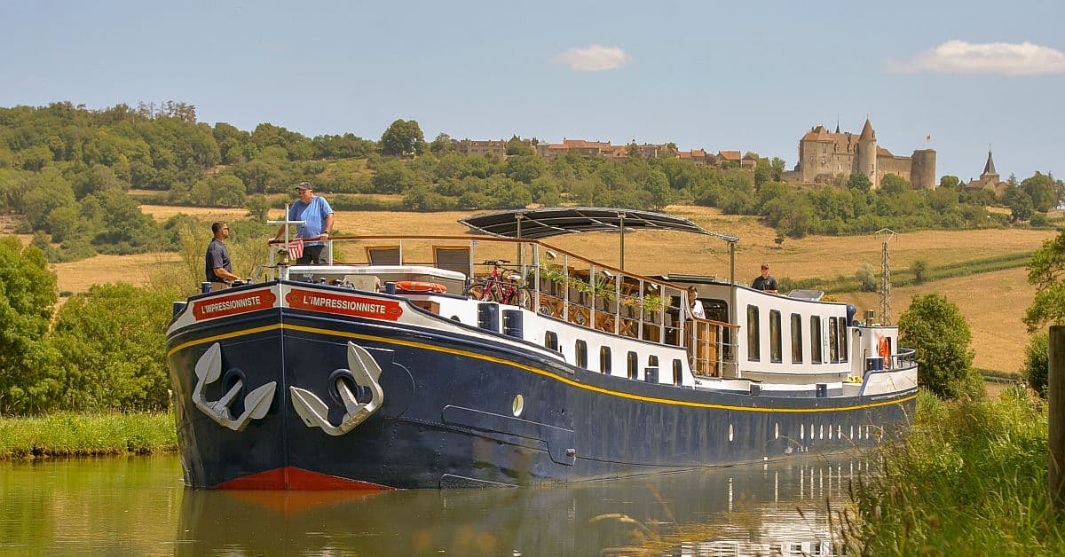 L’Impressionniste Barge on European Waterway A traditional European river barge named L'Impressionniste navigates a peaceful canal with passengers aboard, set against a scenic backdrop of rolling countryside, golden fields, and a medieval castle perched on a distant hilltop.