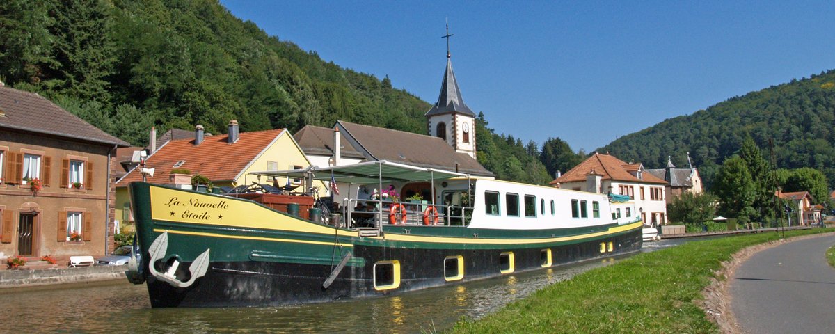 La Nouvelle Étoile Barge in European Canal Village A traditional green and yellow canal barge named 'La Nouvelle Étoile' navigates through a picturesque European village canal, with a charming church steeple and tree-covered hillsides in the background. The barge features typical river cruise accommodations with multiple cabin windows and an outdoor deck area for passengers to enjoy scenic waterway views.