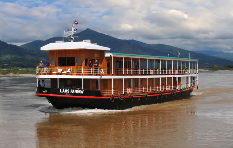 Laos Pandaw River Cruise on Mekong A wooden river cruise boat named Laos Pandaw navigating through a scenic river with mountainous landscape in the background. The multi-deck vessel appears to be traveling on the Mekong River in Southeast Asia.