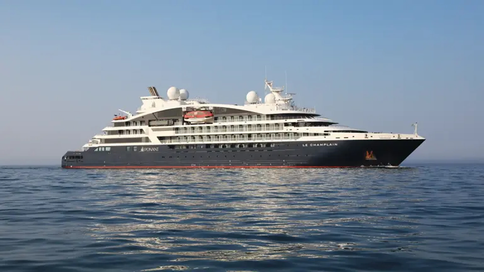 Le Champlain Luxury Expedition Cruise Ship A sleek white and dark gray luxury expedition cruise ship sailing on calm blue waters. The vessel features multiple decks and distinctive white satellite domes on its upper structure.