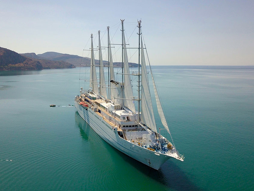 A multi-masted sailing vessel anchored in tranquil blue-green waters with mountainous coastline in the background. The elegant white yacht showcases multiple tall masts and a sleek design against a serene maritime landscape.