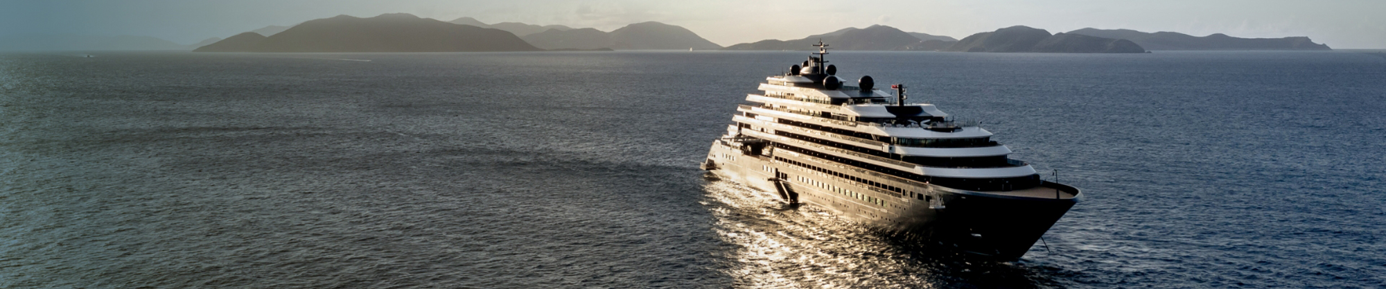 Luxurious Superyacht at Scenic Mountain Coast A sleek, multi-deck white luxury yacht sailing on calm blue waters with a mountainous coastline in the background. The vessel appears to be a high-end private yacht with multiple elegant curved decks.