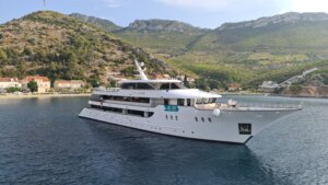 Luxury Cruise Ship Anchored in Mediterranean Bay A modern expedition-style cruise ship is anchored in calm turquoise waters near a picturesque Mediterranean coastal town nestled beneath forested hillsides. The vessel features contemporary white hull design with multiple deck levels and is positioned in a scenic natural harbor.