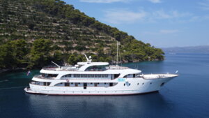 Luxury Cruise Ship Anchored in Mediterranean Waters A multi-deck luxury cruise vessel is anchored in pristine turquoise Mediterranean waters adjacent to a lush, forested mountainous coastline. The white ship features multiple decks and modern amenities, positioned in calm blue waters under clear skies, representing an ideal cruise destination.